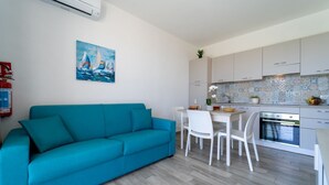 Eine moderne Küche mit weißen Schränken, ein Essbereich mit weißem Tisch und Stühlen sowie ein Salontisch mit einem Türkis-Sofa.