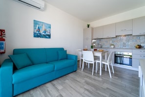 Eine moderne Küche mit weißen Schränken, ein Essbereich mit weißem Tisch und Stühlen sowie ein Salontisch mit einem Türkis-Sofa.