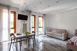 A modern living room with a sofa, a dining table with chairs, a TV mounted on the wall, and large windows with curtains.