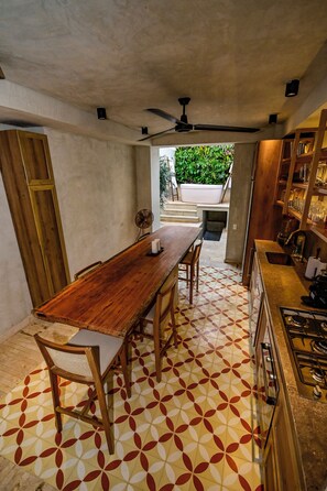 Un espace repas comprenant une table en bois, des chaises et un sol à motifs. On y trouve également un coin cuisine avec une cuisinière et des placards.
