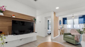 Una sala moderna con un televisor de pantalla plana montado en una pared de madera, un sofá verde, una mesa de centro con flores y vistas al dormitorio contiguo.
