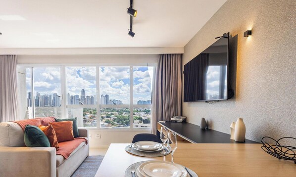 A modern living room with a sofa, dining table, and a city view through large windows. - Metropolitan Sidney - MSS (Goiânia)