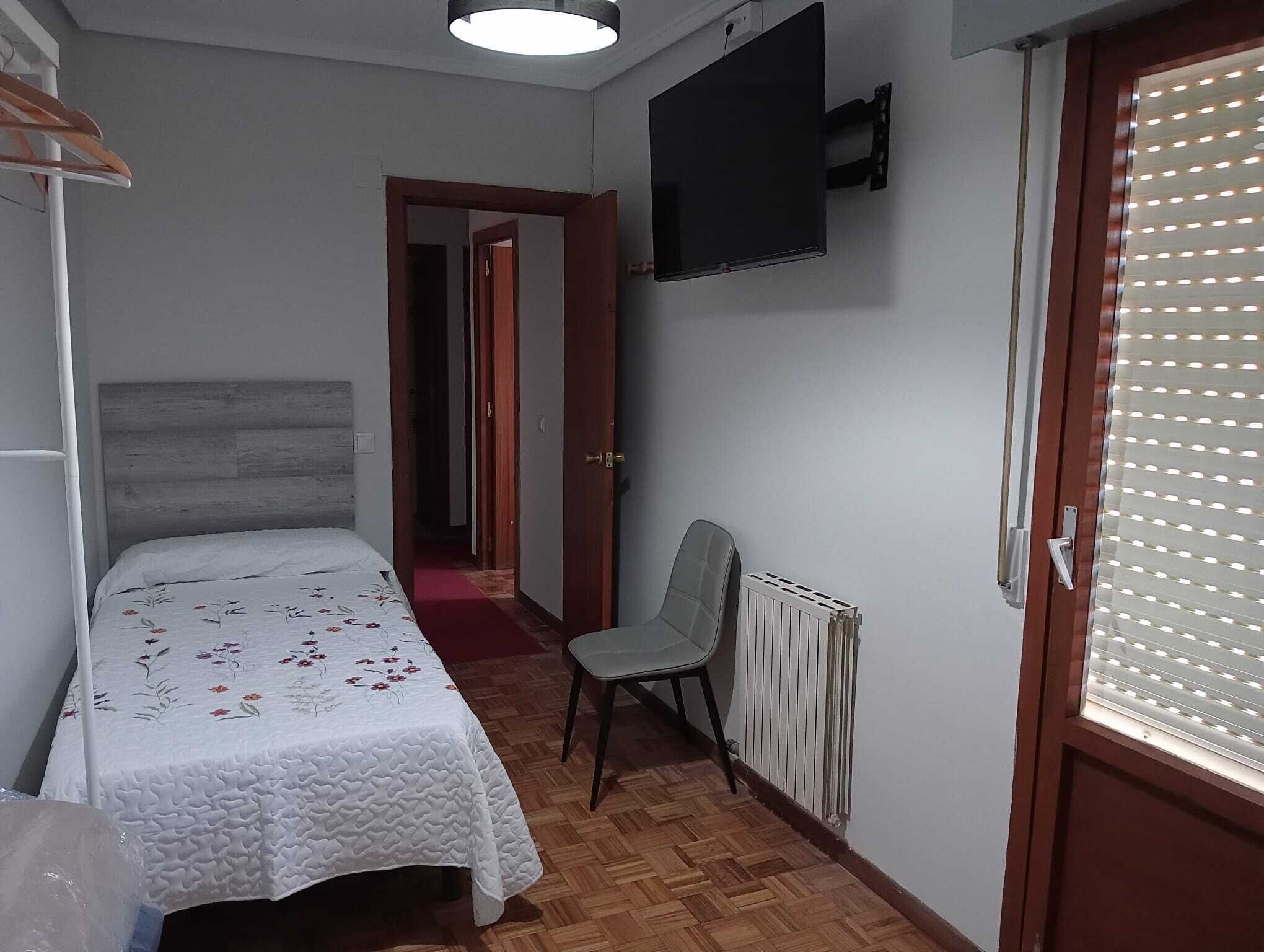Een kleine, verzorgde slaapkamer met een bed, een stoel, een radiator en een aan de muur bevestigde televisie.