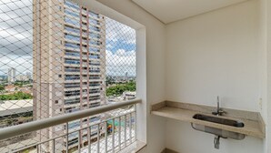 Un balcón con baranda de metal, un lavamanos pequeño y vistas de un paisaje urbano a través de una ventana de malla.