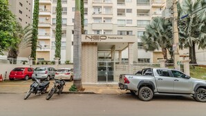 Un edificio de varios pisos con balcones, un letrero que dice 'NEO Practice Home BUENO' y vehículos estacionados, incluyendo una camioneta plateada y motocicletas.