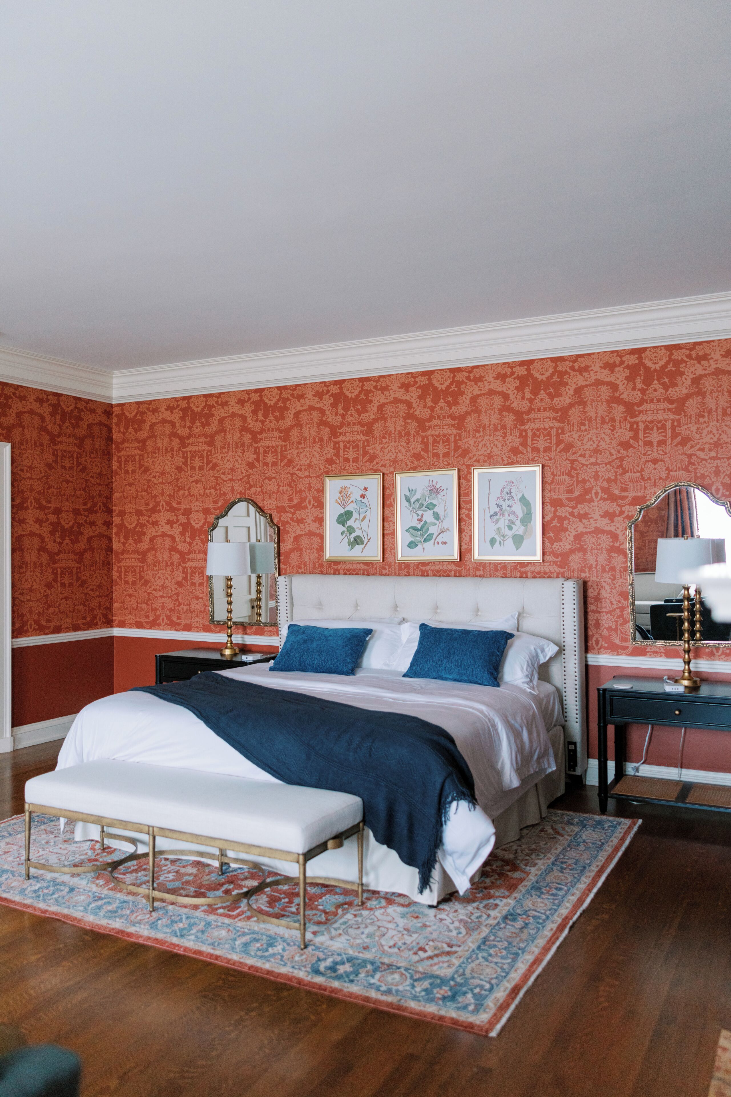 Une chambre à coucher avec un grand lit, un banc, une table de chevet, un miroir et des œuvres d’art encadrées accrochées au mur.