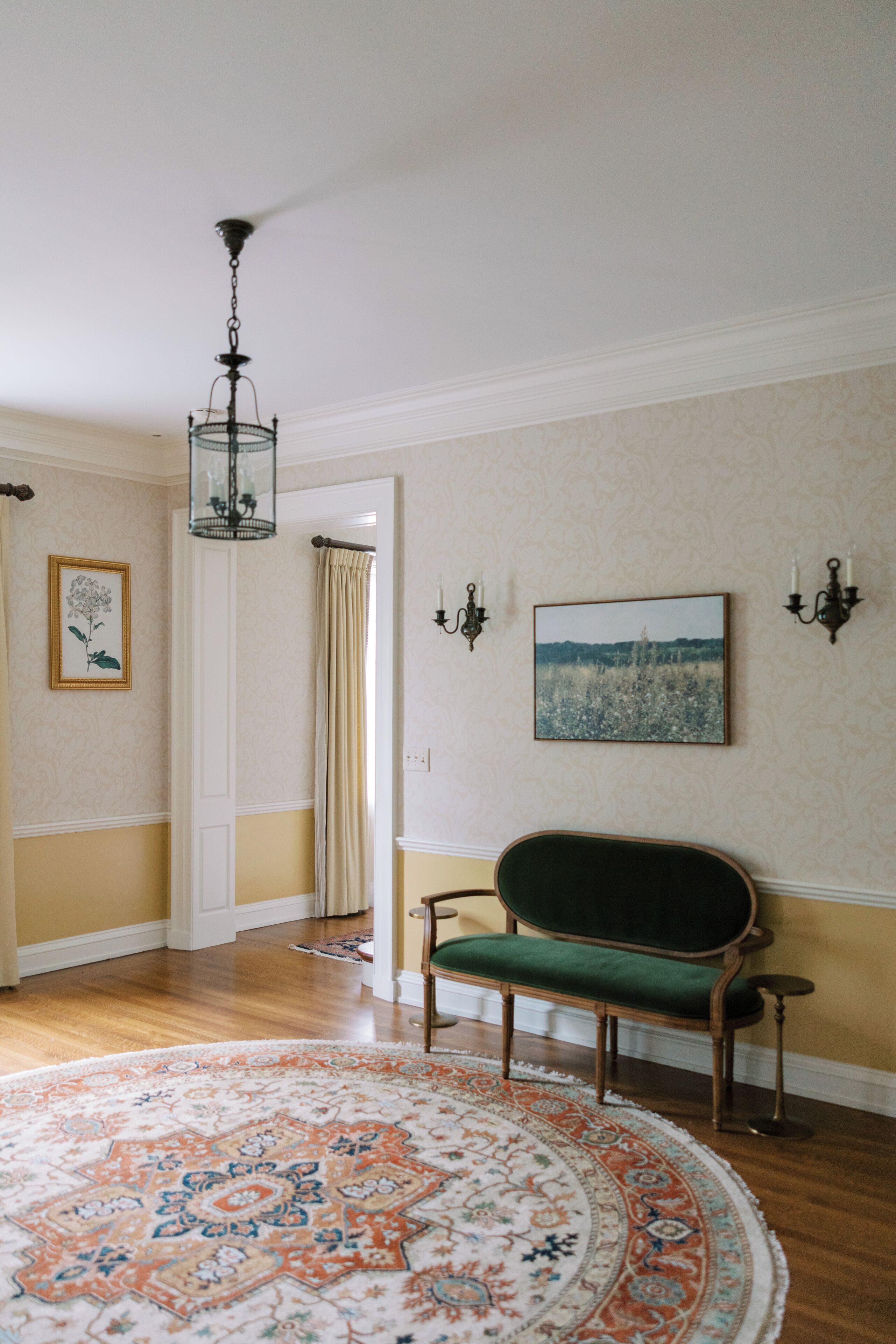 A room with a green upholstered bench, a patterned rug, a chandelier, and wall-mounted artwork.