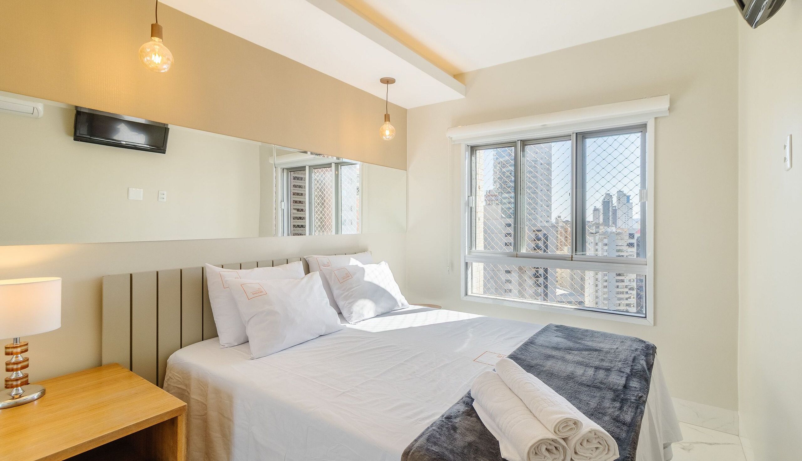 Habitación de hotel con cama, mesita de noche, lámpara, espejo y ventana con vista a la ciudad.