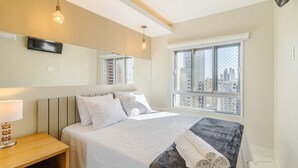 Een hotelkamer met een bed, nachtkastje, lamp, spiegel en een raam met uitzicht op de stad.