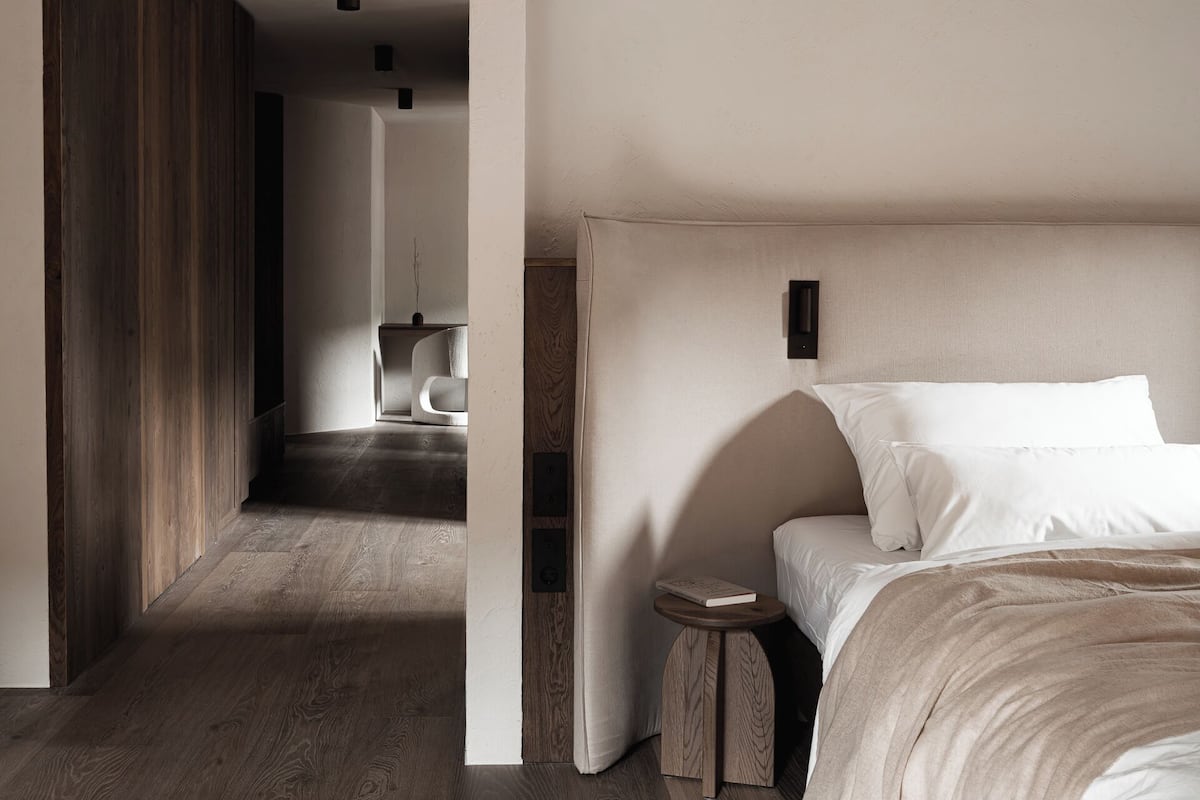 Moderni hotellihuone, jossa on puulattia, suuri sänky valkoisilla vuodevaatteilla, pieni puinen yöpöytä ja näkymä käytävään.