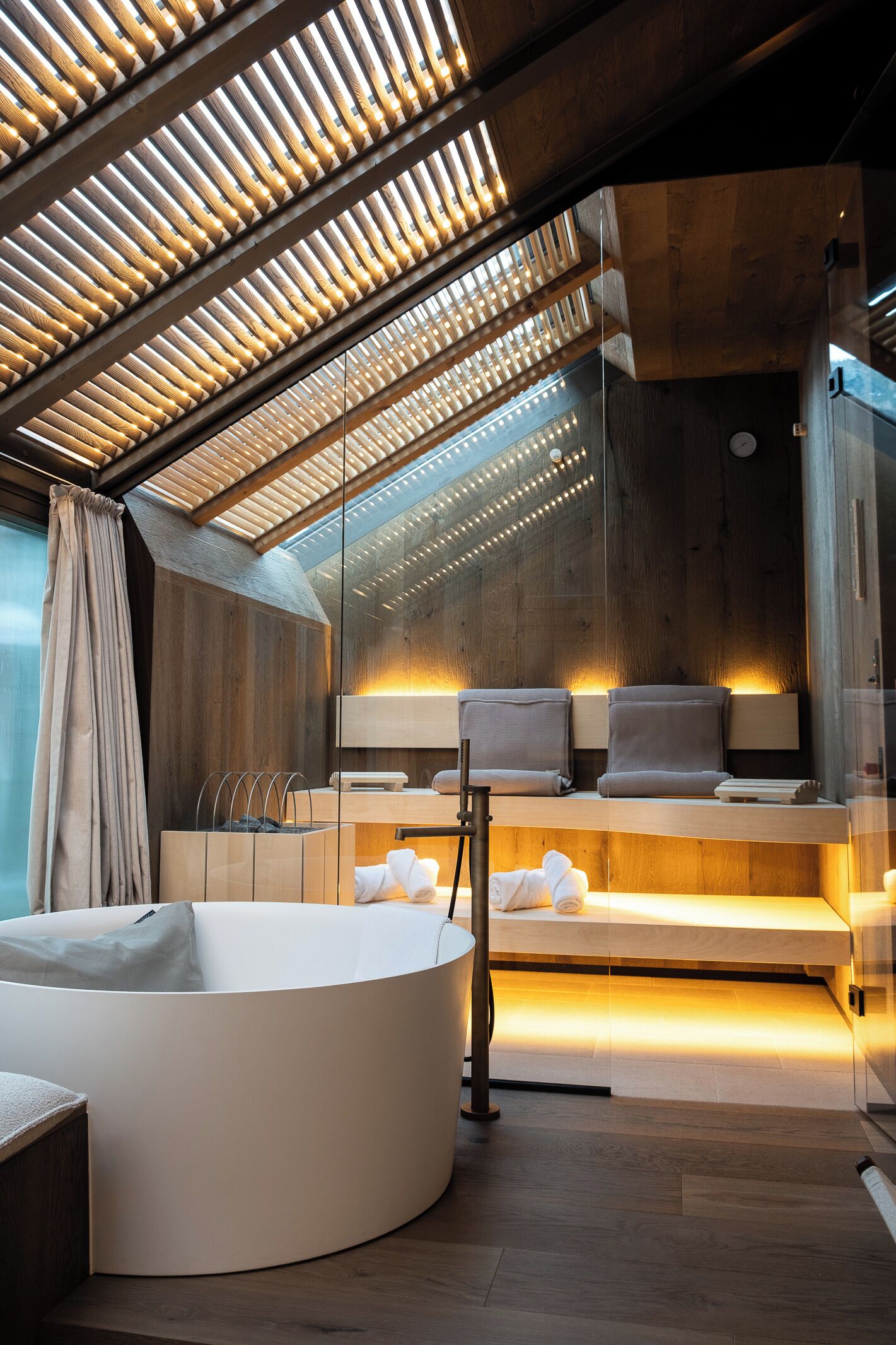 A modern spa room with a large bathtub, wooden ceiling slats, and a bench with towels.