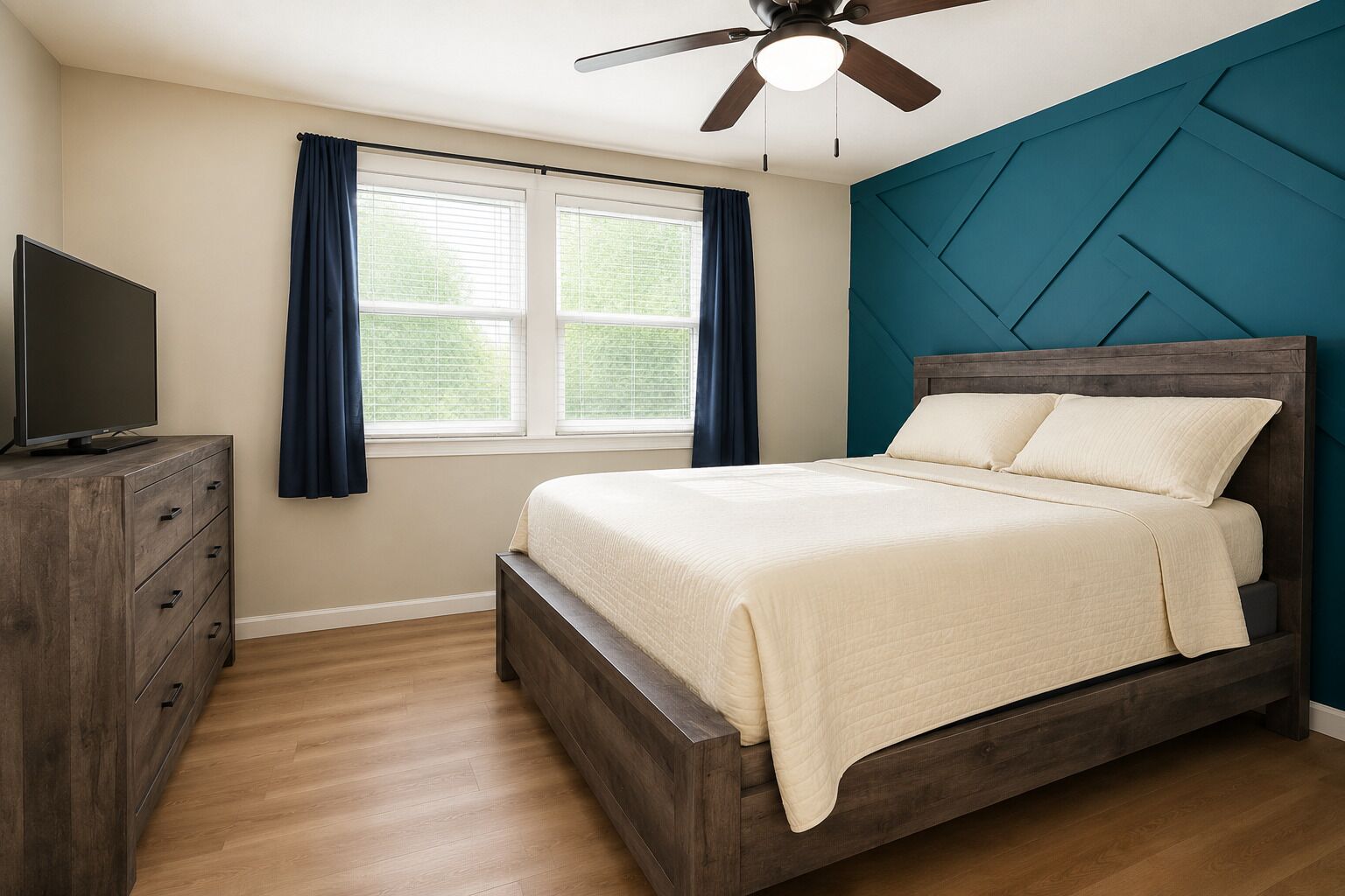 A bedroom with a bed, a TV on a wooden dresser, a ceiling fan, and a window with curtains.
