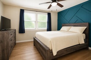 Un dormitorio con cama, televisor sobre un aparador de madera, ventilador de techo y ventana con cortinas.