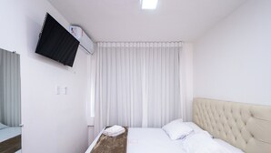 A hotel room with a bed, a flat-screen TV mounted on the wall, and a window with white curtains. - Apt in Full Condo, St. Faicalville - Vam0504 (Goiânia)