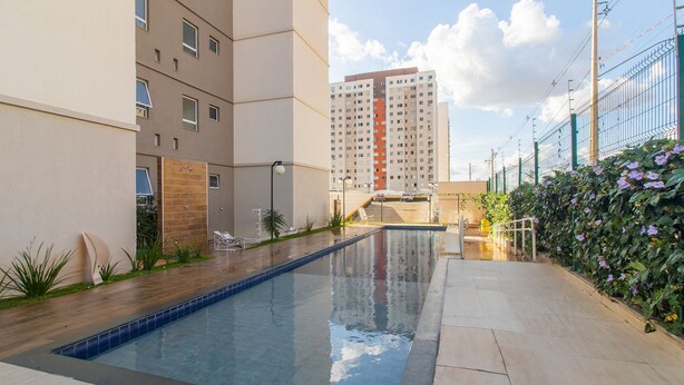 A swimming pool with a tiled deck, a brick wall, and a tall building in the background. - Apt in Full Condo, St. Faicalville - Vam0504 (Goiânia)