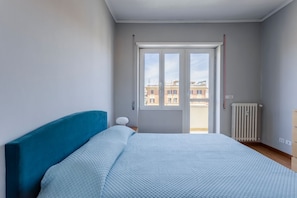 Een slaapkamer met een bed, een raam, een radiator en uitzicht op gebouwen.