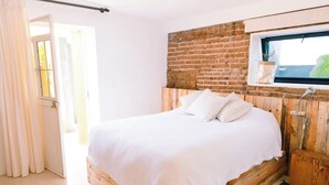 Une chambre avec un lit en bois, du linge de lit blanc, un mur en briques et une fenêtre avec une plante.