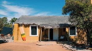 Un bâtiment d’un seul étage, doté d’un toit de chaume, d’un petit porche et d’une grande planche de surf appuyée contre le mur.