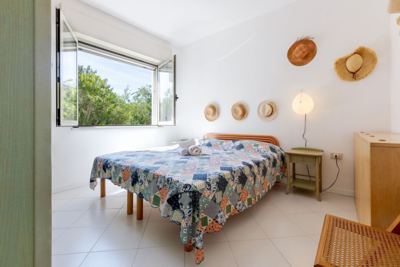 A bedroom with a bed, a window, a bedside table, a lamp, and a wall-mounted hat rack.