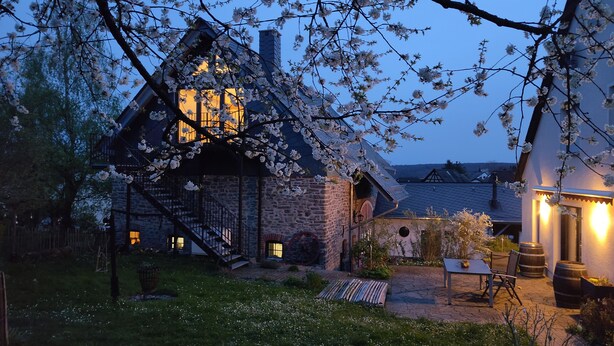 A stone house with a staircase leading to a second floor, surrounded by a garden with white flowers and a paved patio area with outdoor furniture. - "Basalt barn Westerwald" - close to nature & unique (Stahlhofen)