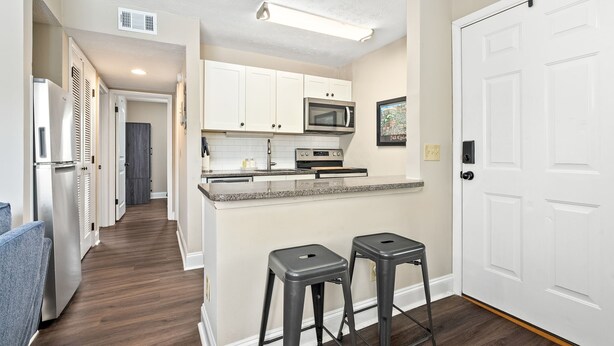 A modern kitchen with white cabinets, a stainless steel refrigerator, a microwave, and a granite countertop. There are two bar stools and a white door to the right.