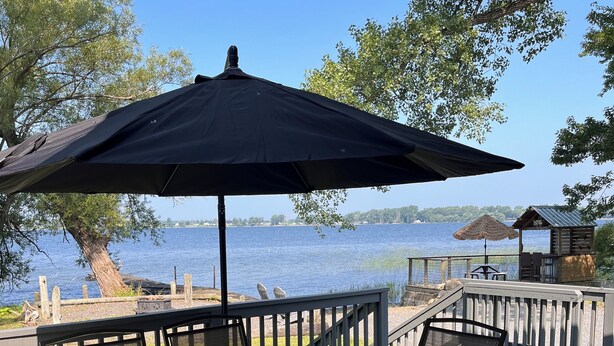 Mesa de vidrio con sillas y un paraguas grande en una terraza de madera con vista a un lago.
