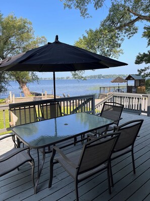 Mesa de vidrio con sillas y un paraguas grande en una terraza de madera con vista a un lago.