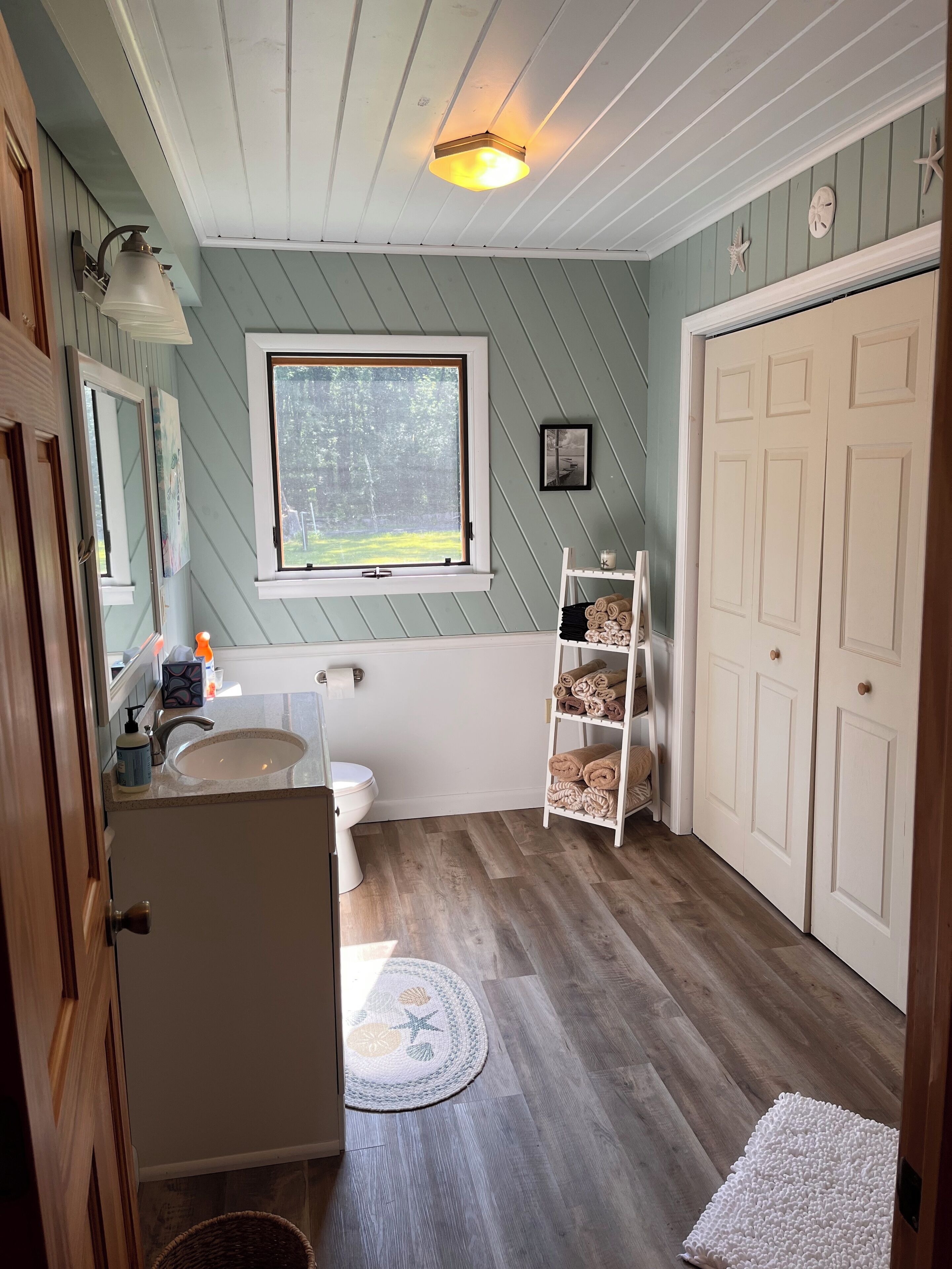 A bathroom with a window, a sink, a toilet, a shelf with towels, and a door.
