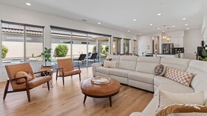 Un salon spacieux avec un grand canapé, deux fauteuils et une table basse. De grandes fenêtres offrent une vue sur une piscine et un espace de détente extérieur.