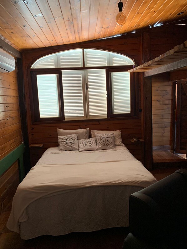 A wooden cabin bedroom with a large bed, a window with blinds, and a hanging light bulb.