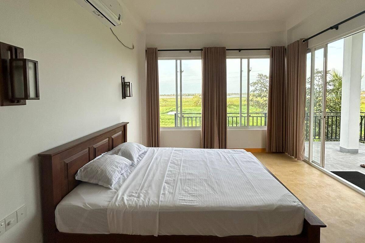 A bedroom with a large bed, an air conditioning unit, a ceiling fan, and a view of a balcony with curtains.
