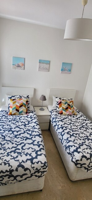 Une chambre avec deux lits, chacun recouvert d’une couvre-lit à motifs, une table de chevet avec une lampe et des œuvres d’art murales représentant des scènes de plage.