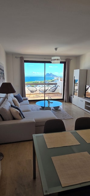 A modern living room with a sofa, a dining table, and a balcony with a view of the sea.