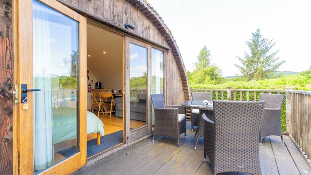 Una terraza de madera con mobiliario de mimbre y un ventanal de vidrio que da a una habitación con cama y mesa.