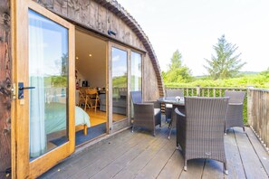 Una terraza de madera con mobiliario de mimbre y un ventanal de vidrio que da a una habitación con cama y mesa.