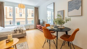 A modern hotel room with a bed, a dining area with a table and chairs, a potted plant, and large windows with a view of buildings. - Designer Prague Studio in a Vibrant Neighborhood by EasyBNB Premium (Prague)