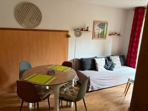 Ein gemütliches Wohnzimmer mit einem runden Esstisch, Stühlen, einer Couch mit Kissen, Holzboden, einem an der Wand befestigten Regal mit Dekoration und einem großen Fenster mit roten Vorhängen.