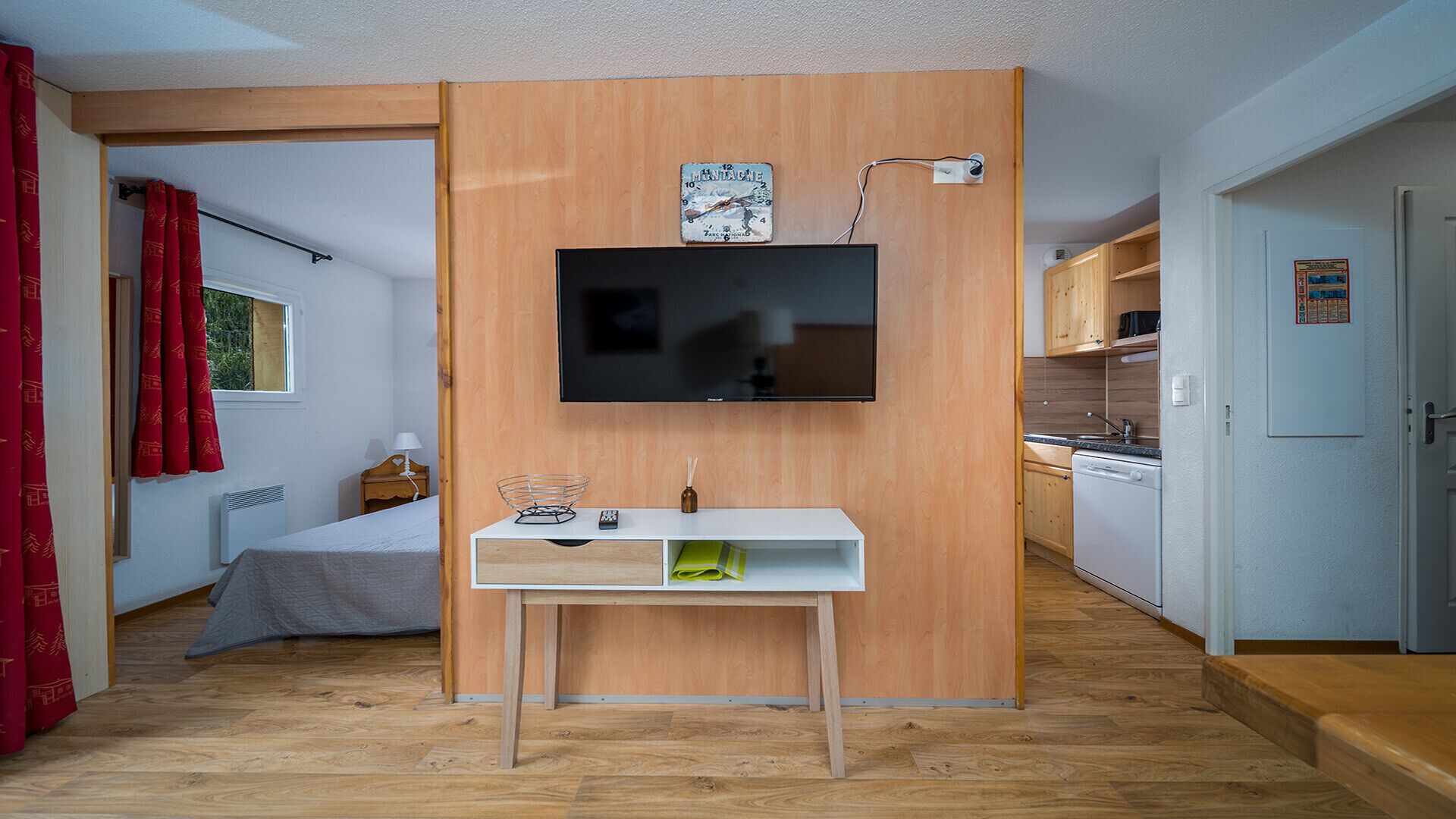 A compact room with a bed, a small desk with a flat-screen TV, a wooden partition, and a kitchenette.