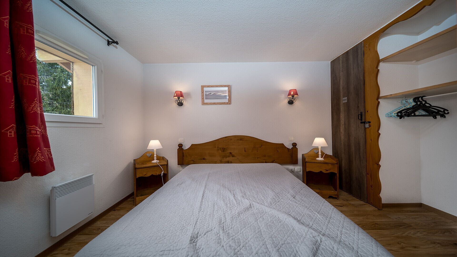 Un dormitorio con cabecera de madera, dos mesitas de noche con lámparas, una ventana con cortinas y un cuadro colgado en la pared.