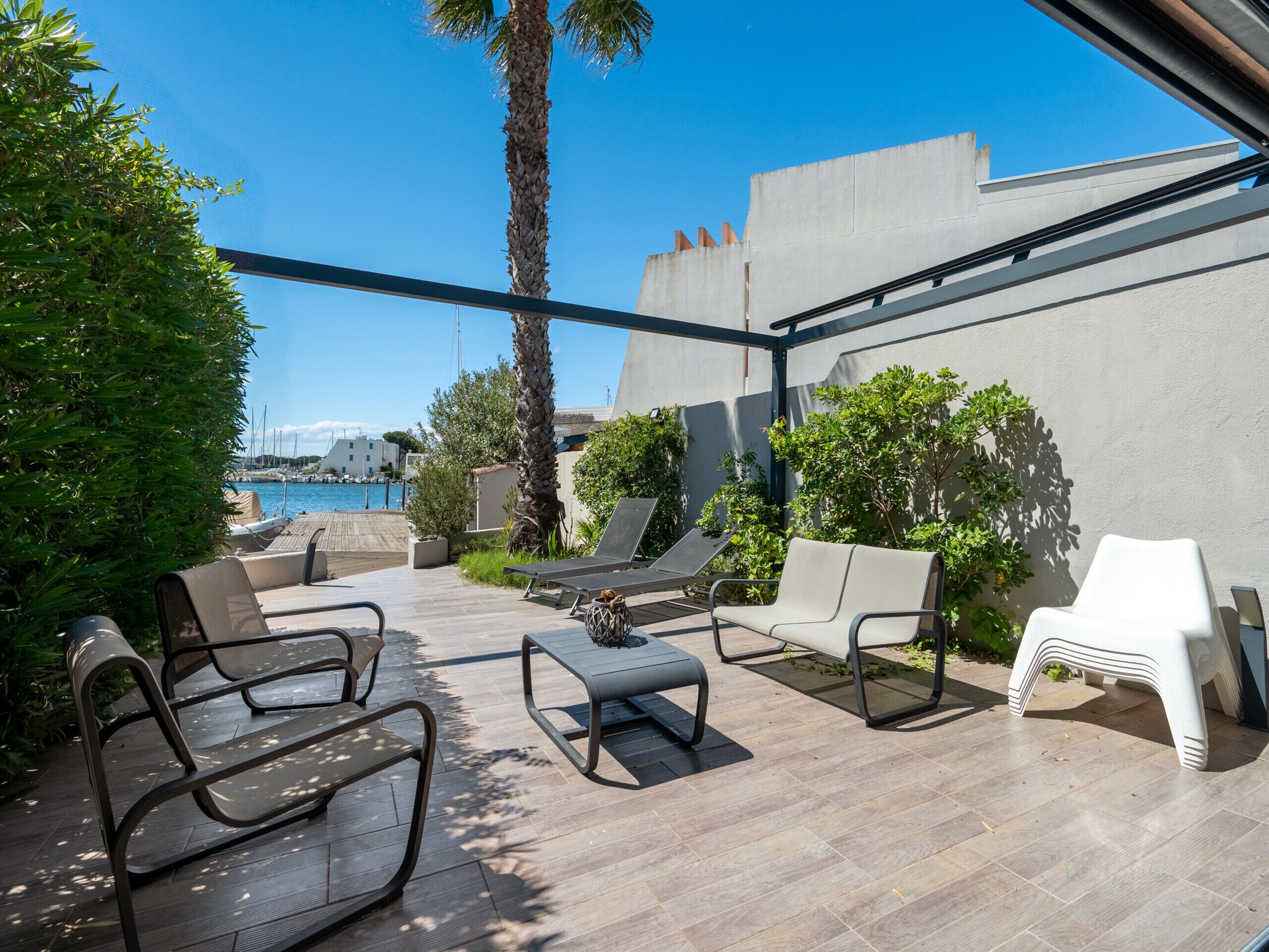 A sunlit outdoor seating area with lounge chairs and a table, overlooking a body of water and a marina.