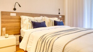 A neatly made bed with striped bedding, a wooden headboard, and a bedside table with a lamp and a small object. - Funchal Acqua Blue 3O, a Home in Madeira (Funchal)