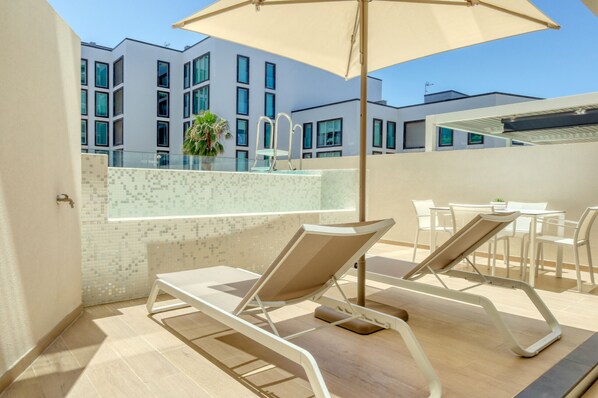 A rooftop terrace with two lounge chairs and a large umbrella, overlooking a modern building with a tiled wall and a pool area.