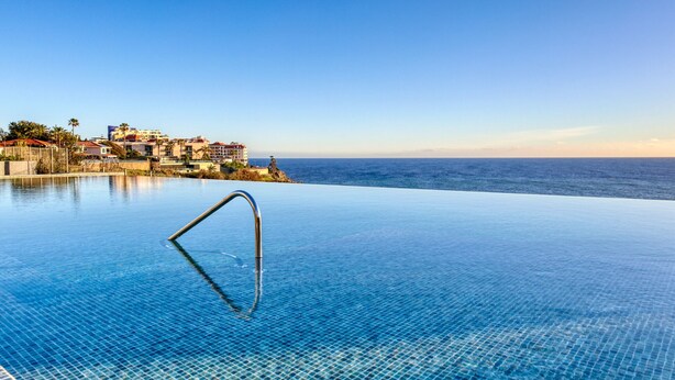 An infinity pool with a clear view of the ocean and a coastal town in the background. - Funchal Acqua Blue 3O, a Home in Madeira (Funchal)