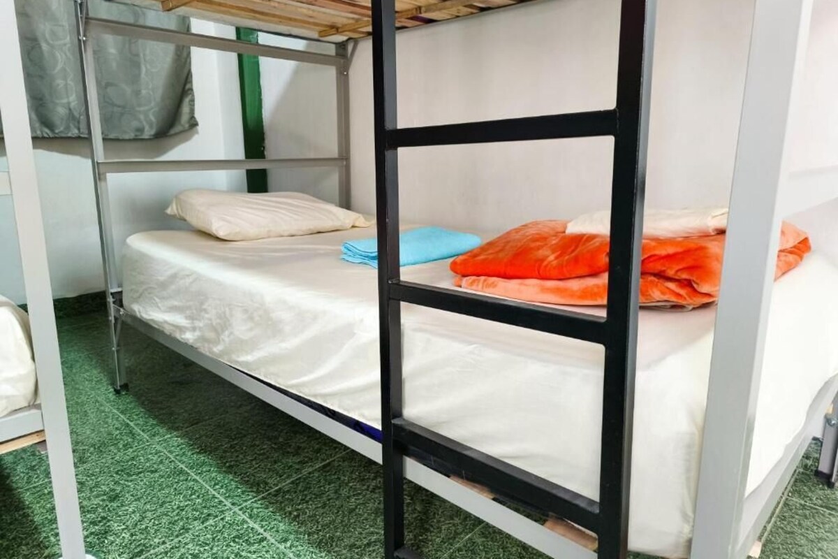 Un lit superposé avec une literie blanche et des oreillers orange, un sol recouvert d’une moquette verte et une structure en métal.