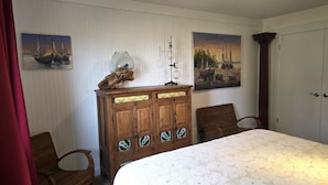 Una habitación con un armario de madera, dos sillas, una cama con colcha blanca y dos cuadros en la pared.