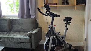 Una habitación con una bicicleta estática, un sofá y una estantería de madera con libros y un bolso.