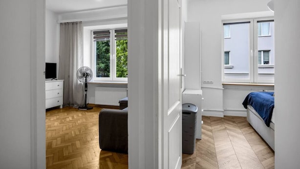 Un dormitorio moderno con piso de madera, una cama con cubierta azul, un ventilador de pie, un radiador y una ventana con vistas a la vegetación.