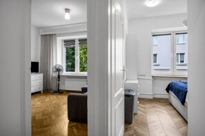 A modern bedroom with a wooden floor, a bed with a blue cover, a standing fan, a radiator, and a window with a view of greenery. - Bokserska Apt - Chopin Airport Nerby - by Rentujemy (Warszawa)