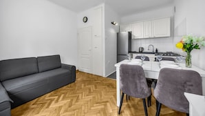 Un espacio habitable compacto con sofá, una mesa de comedor con sillas, una zona de cocina con armarios y electrodomésticos, y un reloj en la pared.