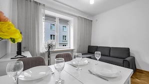 Un área de comedor moderna con una mesa blanca para dos, un sofá gris y una ventana con cortinas.
